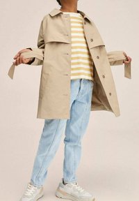 Beżowy trencz z guzikami z przodu, warstwowym krojem i otwartymi rękawami, noszony na pasek w żółto-białe paski, w połączeniu z jasnoniebieskimi dżinsami i trampkami.