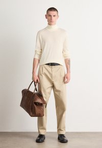 Pull en col roulé beige, couleur crème, associé à un pantalon beige. Sac de voyage en cuir marron, ceinture noire et chaussures noires. Design simple.