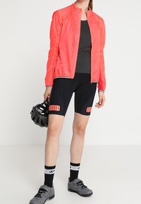 Světlá růžová lehká cyklistická bunda, šedý strukturovaný upnutý top, černé cyklistické kraťasy s růžovými akcenty, šedé sportovní boty a černá helma.