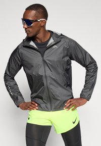 Letti sort jakke med reflekterende accenter og en mønstret hætte. Båret over en sort t-shirt og lyse neon-grønne shorts. Mørke solbriller.