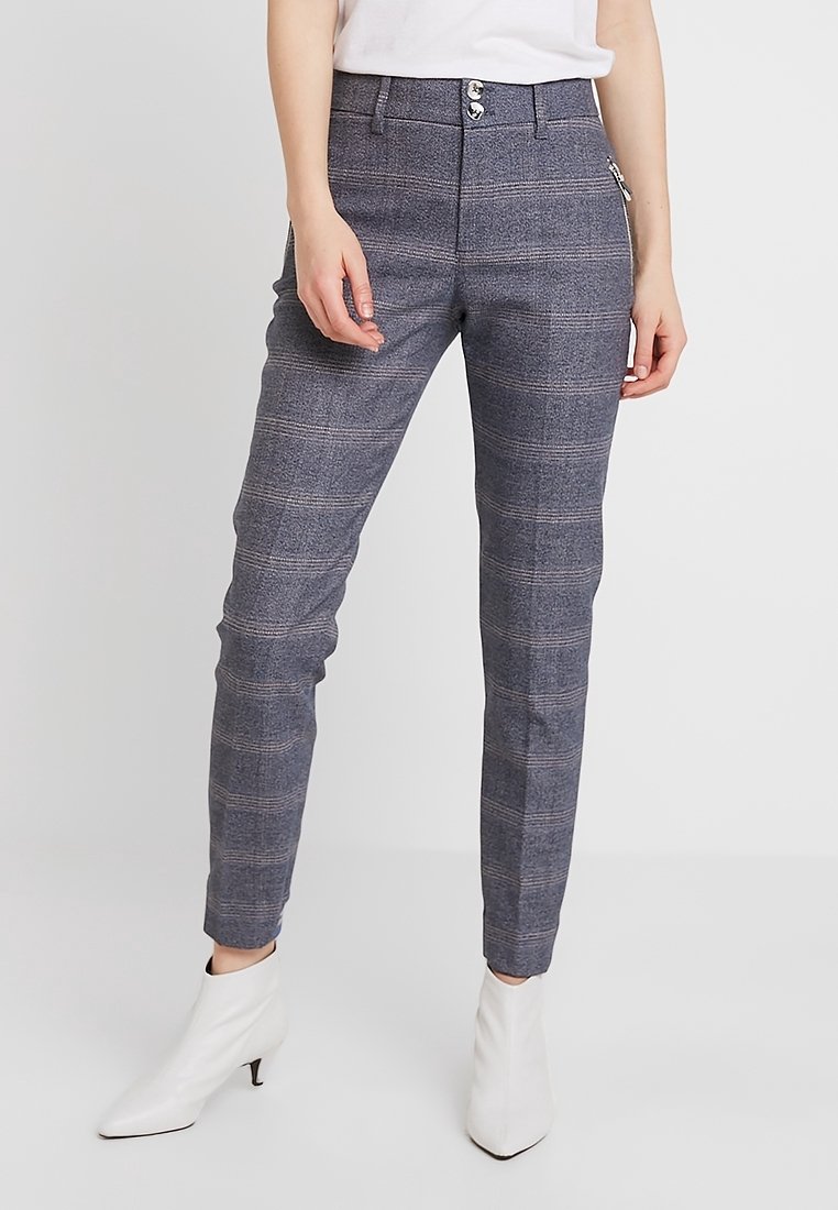Pantalons à carreaux gris avec une coupe ajustée, présentant une fermeture éclair, deux poches avant et une texture subtile. Portés avec des bottines blanches.