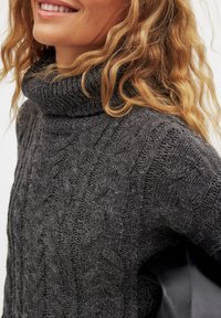 Grå turtlenecktröja med kabelstickad mönster, tjock textur och rullad halsringning. Svart läderdetalj synlig på sidan.