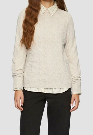 Femme aux longs cheveux bruns portant un pull crème en maille torsadée par-dessus une chemise blanche à rayures noires et un pantalon noir.