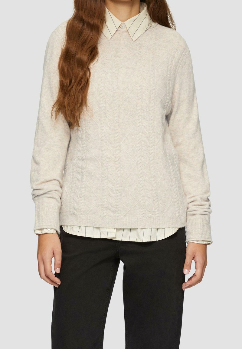 Femme aux longs cheveux bruns portant un pull crème en maille torsadée par-dessus une chemise blanche à rayures noires et un pantalon noir.
