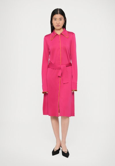 Różowa sukienka z długim rękawem, kołnierzykiem, zapięciem na guziki z przodu, paskiem w talii oraz żółtymi lamówkami na kołnierzyku i mankietach. Gładka faktura materiału.