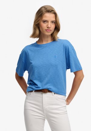 Giovane donna con capelli biondi fino alle spalle, che indossa una t-shirt blu a maniche corte infilata in pantaloni bianchi con una cintura intrecciata marrone.