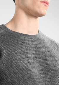 Pull en maille texturée grise avec un col rond. Le tissu semble tricoté, présentant un motif subtil et une coupe ajustée au niveau des épaules.