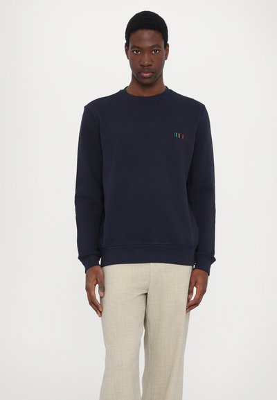 Sweatshirt bleu marin avec un col rond et des poignets côtelés, présentant un petit motif de rayures colorées sur la poitrine. Porté avec un pantalon de couleur claire.
