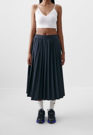 Femme portant un top court blanc, une jupe midi plissée noire, des chaussettes blanches Nike et des baskets noires à semelles bleues, posant devant un fond uni.