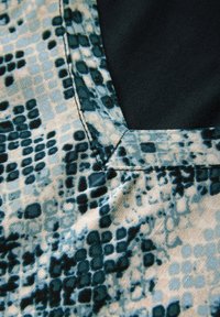 Textil mit einem blau-schwarzen Schlangenhautmuster, auf einer Seite mit einem kontrastierenden schwarzen Stoff und sichtbaren genähten Kanten.