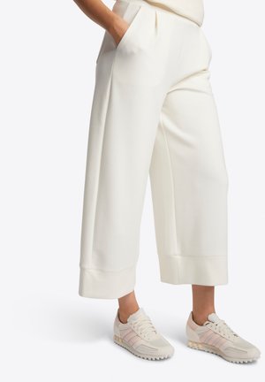 Femme portant un pantalon blanc court et ample avec des poches et des baskets blanches avec des rayures rose pâle, debout avec une main dans la poche.