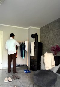 Man organizing clothes on wooden rack in modern room with black stove, gray ottoman, and patterned armchair with folded blankets.