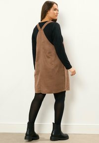 Vestido marrón de ante con tirantes ajustables y corte recto, combinado con una camisa negra de manga larga y botas negras hasta el tobillo.