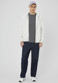 Sweat à zip gris clair, t-shirt gris foncé, jean bleu foncé ample, et baskets blanches, surmontés d'une casquette violet clair.