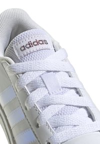 White athletic shoe with synthetic upper, featuring textured mesh tongue, flat white laces, and three striped accents in a lighter shade.