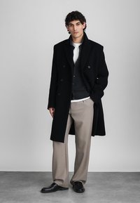 Homme debout portant un manteau croisé noir, un cardigan gris foncé, une chemise blanche, un pantalon beige et des chaussures de ville noires, sur un fond gris clair.