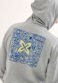 Sudadera gris con un diseño gráfico azul que presenta varias formas abstractas y un elemento floral amarillo en la parte posterior. Textura suave, corte holgado.