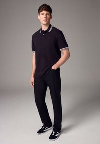 Fekete póló, csíkozott gallérral, fekete nadrággal és fehér részletekkel ellátott fekete sportcipőkkel párosítva. A modellen az egyik kezében zsebében pózol.