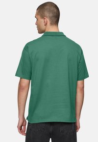 Grön bomullspolo skjorta sedd bakifrån, med klassisk krage och korta ärmar i en avslappnad passform, tillsammans med mörka byxor.