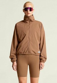 Braune, leichte Jacke mit hohem Kragen und Reißverschluss, kombiniert mit passenden Shorts. Reflexion Sonnenbrille mit pinken Gläsern.