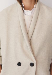 Manteau beige croisé en mélange de laine, avec une finition satinée, de gros boutons noirs et un col châle porté sur un chemisier blanc à volants.