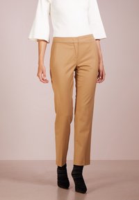 Kvinna i beige skräddarsydda byxor, vit topp med trekvartsärm och svarta rynkade ankelstövlar som står mot en neutral bakgrund.