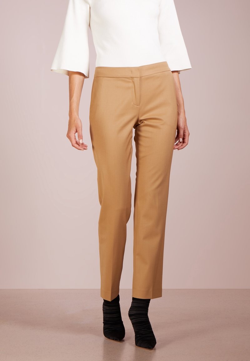 Kvinna i beige skräddarsydda byxor, vit topp med trekvartsärm och svarta rynkade ankelstövlar som står mot en neutral bakgrund.