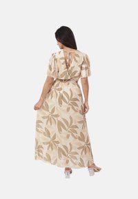 Mujer de pie con la espalda vuelta, vestida con un maxi vestido floral beige con mangas abullonadas y sandalias de tacón bloque transparente.