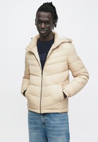 Μπεζ μπουφάν puffer με φερμουάρ μπροστά, κουκούλα και ραμμένες λεπτομέρειες. Φορεμένο πάνω από ένα μπλε πουλόβερ με σχέδια και συνδυασμένο με μπλε τζιν.