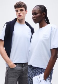 Νεαρός άνδρας και γυναίκα φοράνε λευκά T-shirts Lacoste· ο άνδρας έχει μπλε σκούρο πουλόβερ περασμένο πάνω από τους ώμους, η γυναίκα κρατάει λευκό ρούχο με μπλε περιθώριο.