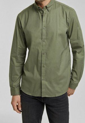 Mężczyzna w oliwkowej, długorękawowej koszuli na guziki z kołnierzykiem, ubrany w czarne jeansy, stoi na tle jednolitego szarego tła.