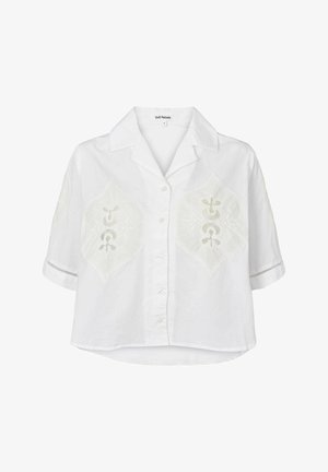 Camicia bianca a maniche corte con bottone frontale, caratterizzata da motivi a rombi ricamati sul petto e cuciture decorative sulle maniche.