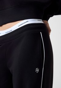 Czarne sportowe spodnie z białym paskiem z logo z napisem "GUESSJEANS", białym lamowaniem po bokach oraz miękką, gładką teksturą.