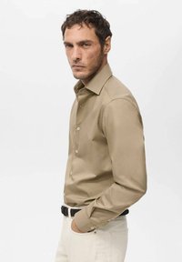 Camisa de manga larga beis con cuello abotonado, fabricada en material suave y de corte estándar. Presenta botones en la parte delantera y un acabado pulido.