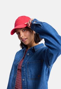 Rote Baumwollmütze mit verstellbarem Riemen, getragen zu einem blauen Jeanshemd mit Tasche und Knopfdetails sowie einem gestreiften rot-weißen Oberteil darunter.