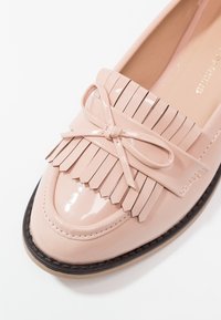 Mocassins en cuir verni rose blush avec détails à franges, accent nœud, et semelle en caoutchouc noire. Texture lisse et design à bout arrondi.