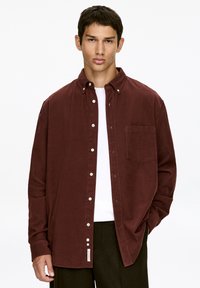 Bordeauxfarbenes Hemd aus Cordstoff mit einer Brusttasche, weißen Knöpfen und einer lockeren Passform. Über einem weißen T-Shirt getragen.