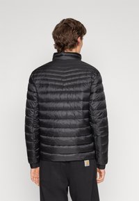 Osoba otočená zády, oblečená v černé prošívané péřovce a černých kalhotách na jednobarevném světlém pozadí.