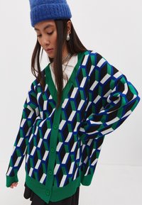 Pestrý pletený cardigan s geometrickým vzorem v modré, zelené, černé a bílé barvě; obsahuje zelený žebrovaný límec a knoflíky.