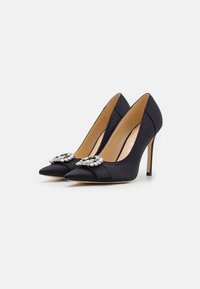Tacchi alti in satin nero con punta appuntita, caratterizzati da un dettaglio con una spilla di gioiello sulla tomaia e un sottile tacco a stiletto.
