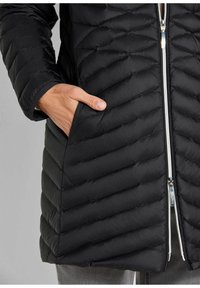 Zwarte pufferjas met een zigzag quiltpatroon, voorrits en zijzakken. Het materiaal lijkt lichtgewicht en glad.