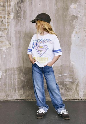 Ung pige med langt blondt hår, iført en "Supergirl" grafisk t-shirt, baggy jeans og en sort kasket, står op ad en struktureret væg.