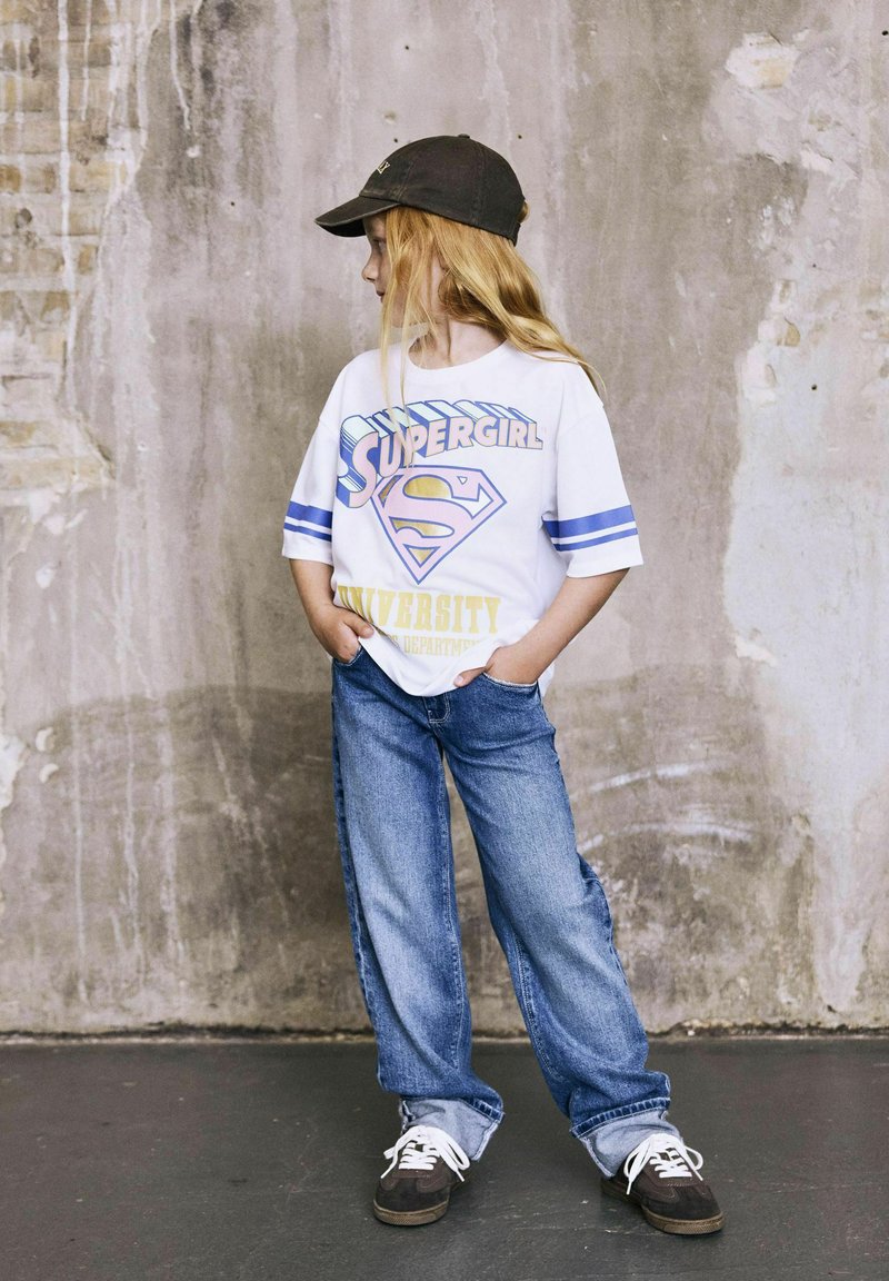 Jeune fille aux longs cheveux blonds, portant un t-shirt graphique "Supergirl", un jean ample et une casquette noire, se tient devant un mur texturé.
