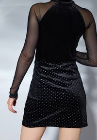 Bodysuit en velours noir avec des manches longues transparentes, associé à une jupe noire ornée de clous en argent, présentant une coupe ajustée et un léger brillant.