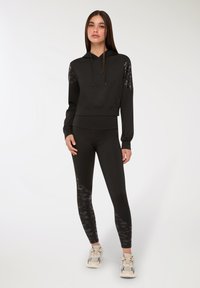 Donna in piedi con un felpa nera con cappuccio e leggings neri con pannelli a fantasia, scarpe da ginnastica chiare, sfondo neutro.