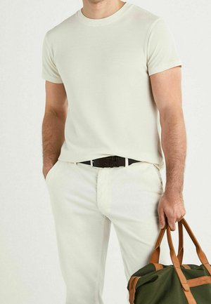 T-shirt côtelé crème associé à un pantalon beige clair. Ceinture marron en détail, tenant un sac vert olive avec des accents en cuir marron clair.