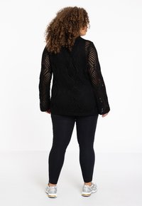 Zwarte gebreide trui met een open netontwerp, lange mouwen en een ronde hals, gecombineerd met zwarte leggings en zilveren sportschoenen.