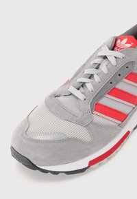 Grijze suede en mesh sneaker met rode en zilveren accenten, voorzien van een ronde neus, gevoerde tong en grijze veters. Rubberen zool zichtbaar.