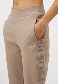 Beige sweatpants med en glat tekstur, elastisk talje, sidelommer og dekorativ broderi langs siderne, med "GUESS" logo.