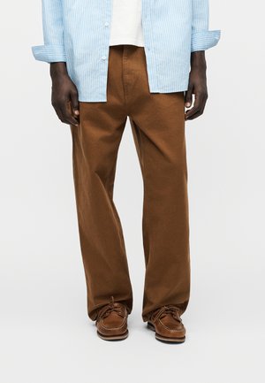 Moški, ki nosi ohlapne rjave hlače, rjave čolnarske čevlje, belo majico in modro-belo črtasto srajco z zavihanimi rokavi.